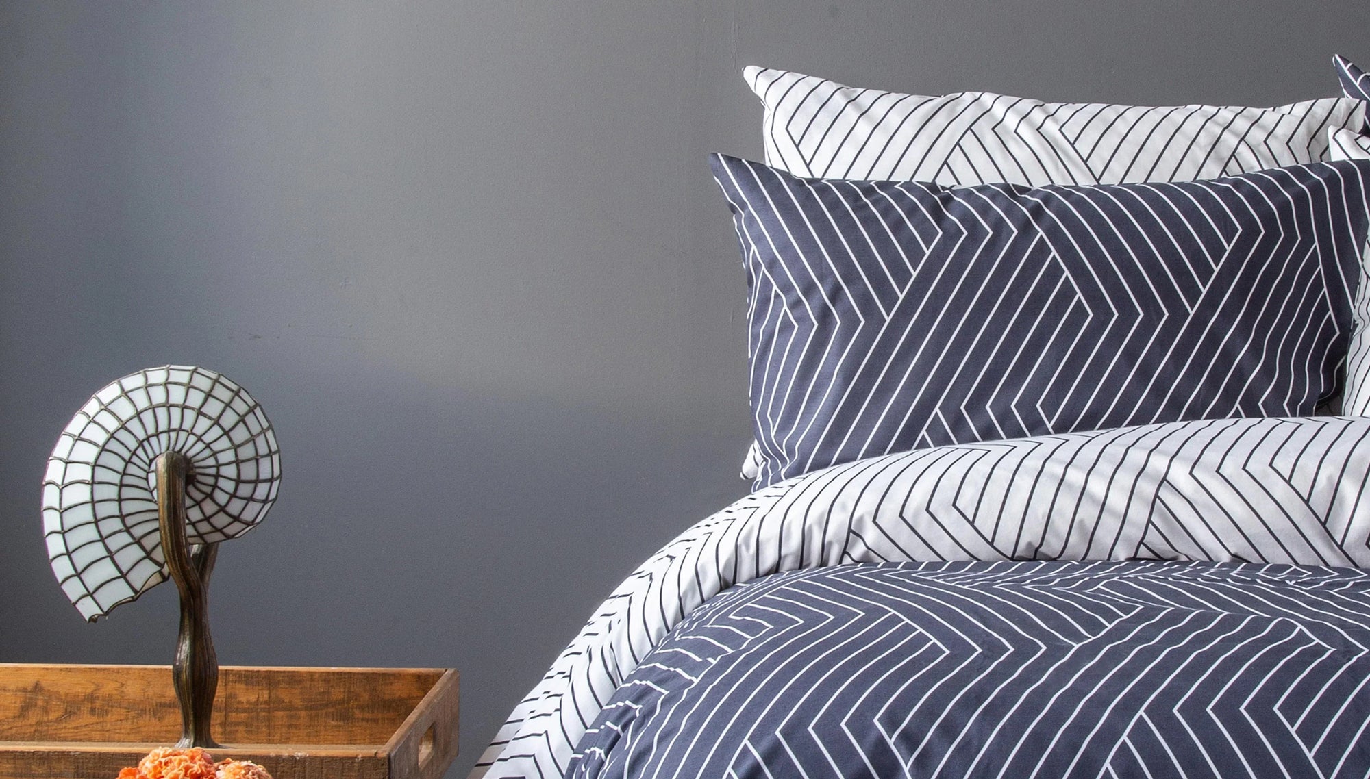 Modern navy and white striped bedding set with geometric patterns, beside a wooden nightstand and decorative lamp.