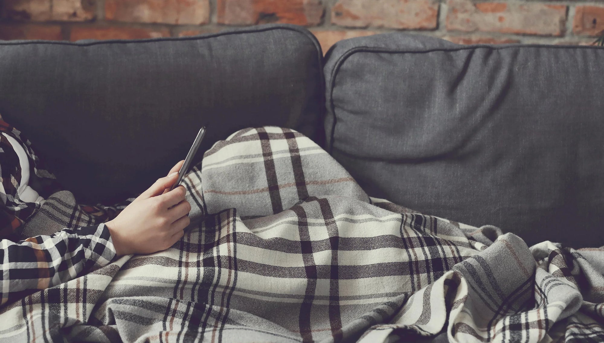 Person using smartphone on grey sofa with plaid blanket and brick wall background